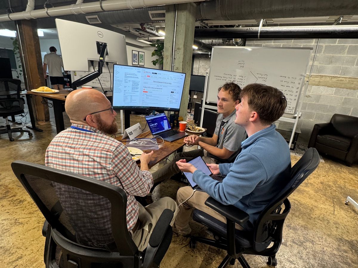A mentor sits with two students at a desk with a large monitor.