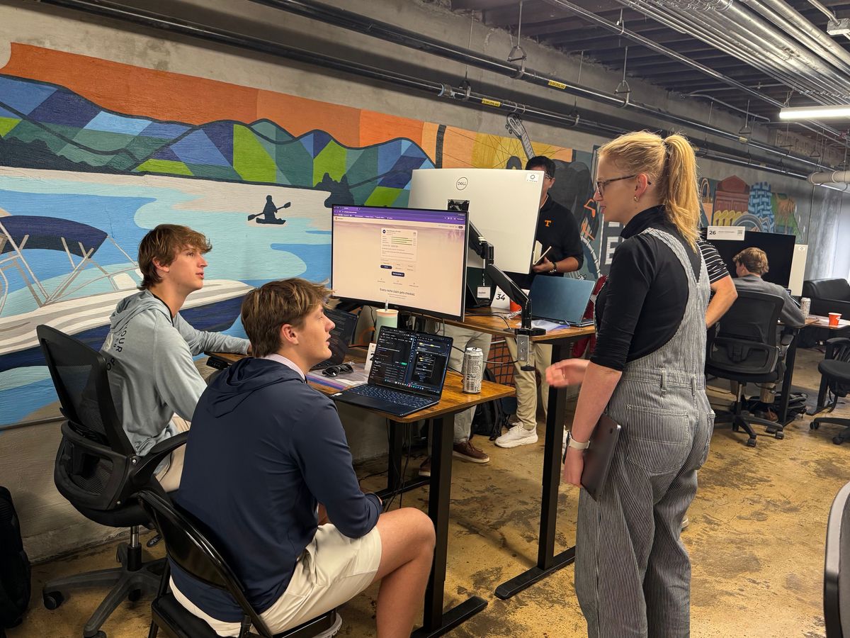 A mentor stands with two students working at a monitor in front of a mural.